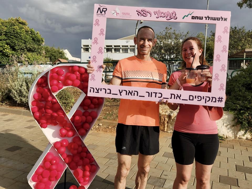 גבר ואישה משתתפים במרוץ להעלאת מודעות לסרטן השד בדרום השרון ומחזיקים שלט עם הכיתוב גם אני רצה מקיפים את כדור הארץ בריצה